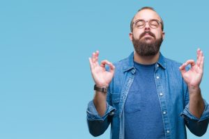 Young caucasian hipster man wearing glasses over isolated background relax and smiling with eyes closed doing meditation gesture with fingers. Yoga concept.